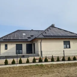 Jednopiętrowy dom z beżową elewacją i ciemną dachówką, na której zamontowane są panele fotowoltaiczne oraz okna dachowe, ogrodzony metalową siatką i obsadzony tujami.