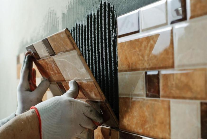 Układanie brązowo-beżowych płytek ceramicznych w kształcie cegiełek na ścianie z użyciem czarnego kleju, widoczne ręce w białych rękawicach roboczych z pomarańczowym wykończeniem.