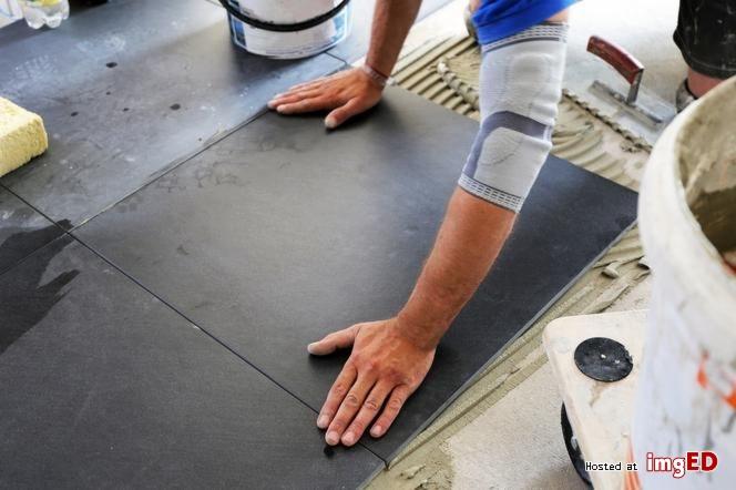 Układanie ciemnych, matowych płytek podłogowych; widoczna dłoń glazurnika w stabilizatorze na kolanie, wiadro zaprawy i packa do rozprowadzania kleju.