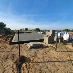 Wylana płyta fundamentowa z izolacją, przygotowana pod budowę domu; na placu budowy palety z bloczkami betonowymi i materiałami izolacyjnymi.