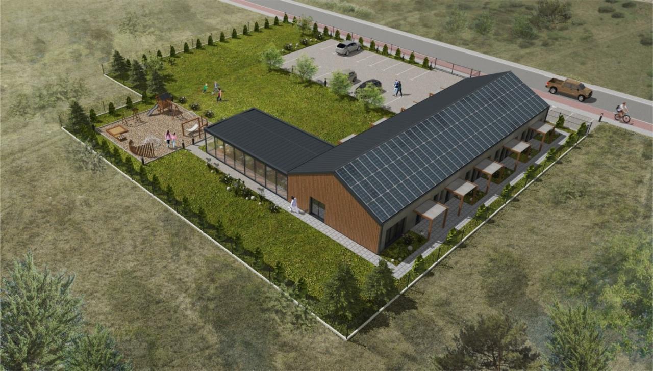 Wizualizacja z lotu ptaka nowoczesnego budynku z panelami słonecznymi na dachu, przyległym parkingiem, placem zabaw i zagospodarowanym terenem zielonym.