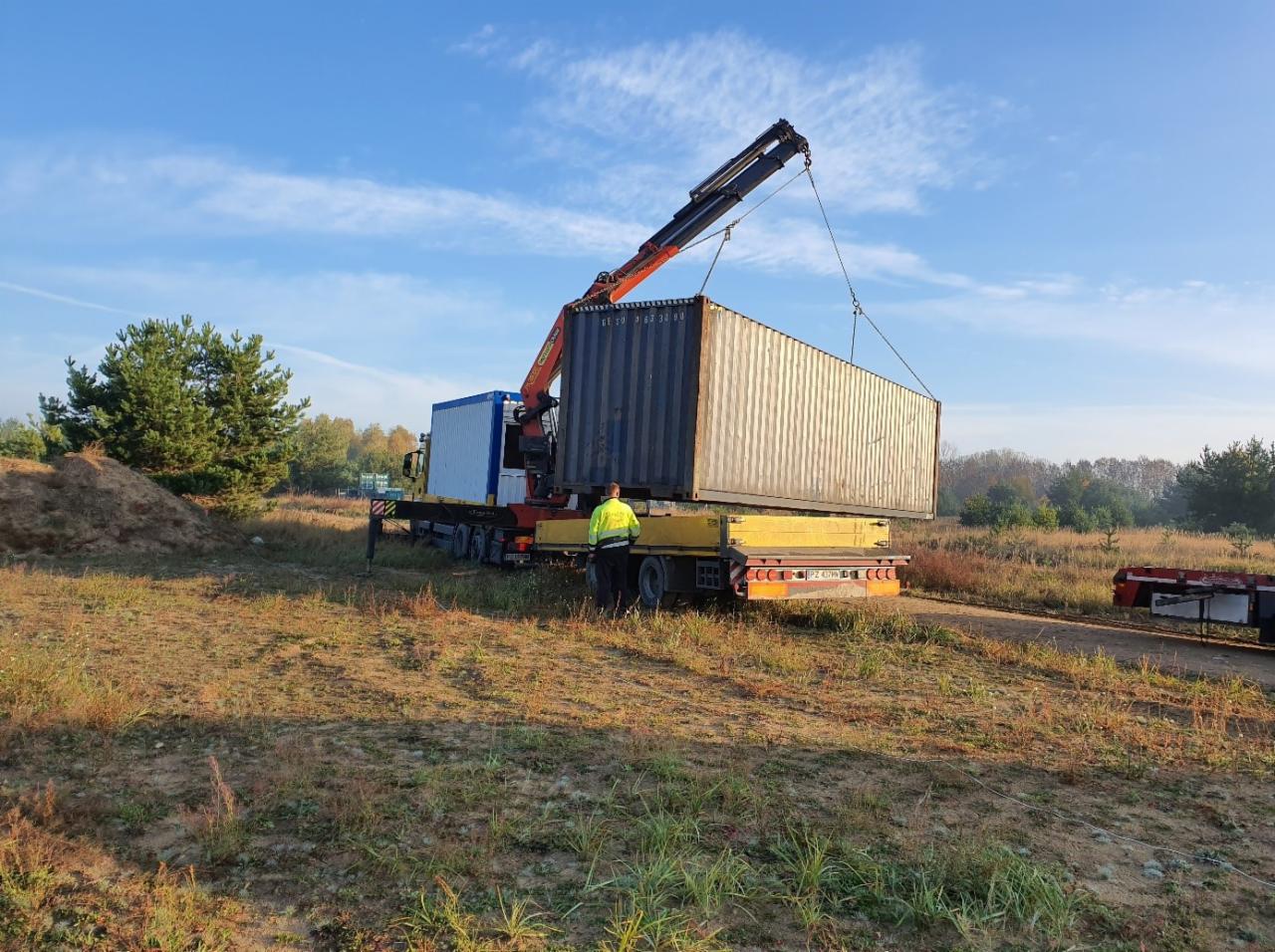 Rozładunek kontenera morskiego za pomocą dźwigu samochodowego na przyczepę niskopodwoziową, operator w żółtej kamizelce asekuruje operację.