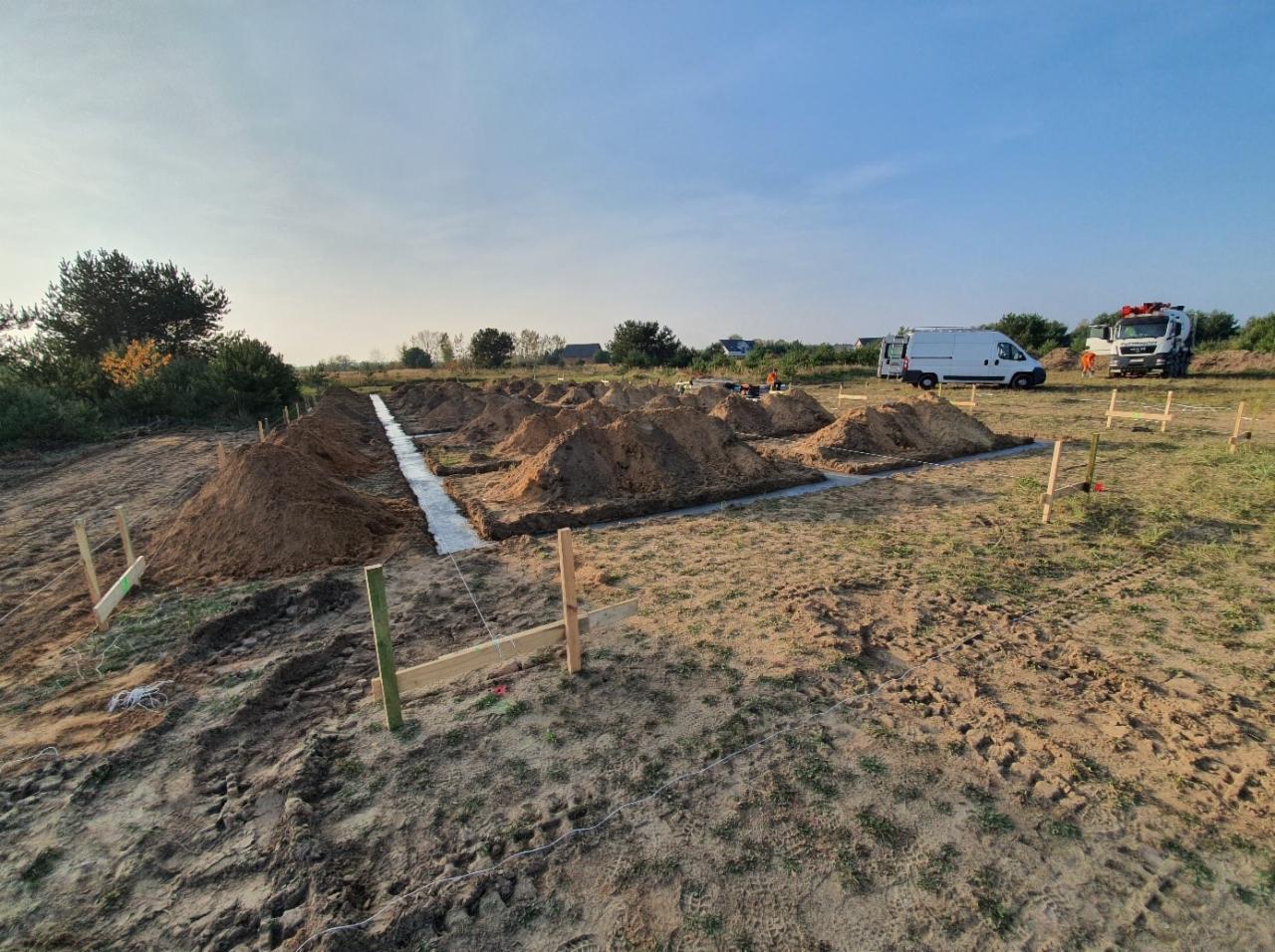 Przygotowanie terenu pod budowę: wykopane fundamenty, częściowo zalane betonem, usypane kopce ziemi, wbite drewniane paliki z linkami wyznaczającymi granice, na dalszym planie samochód dostawczy...