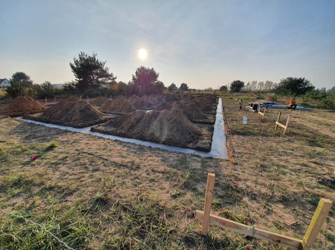 Wykonane wykopy pod fundamenty domu jednorodzinnego, zalane betonem, widoczne pryzmy ziemi i drewniane elementy szalunku.