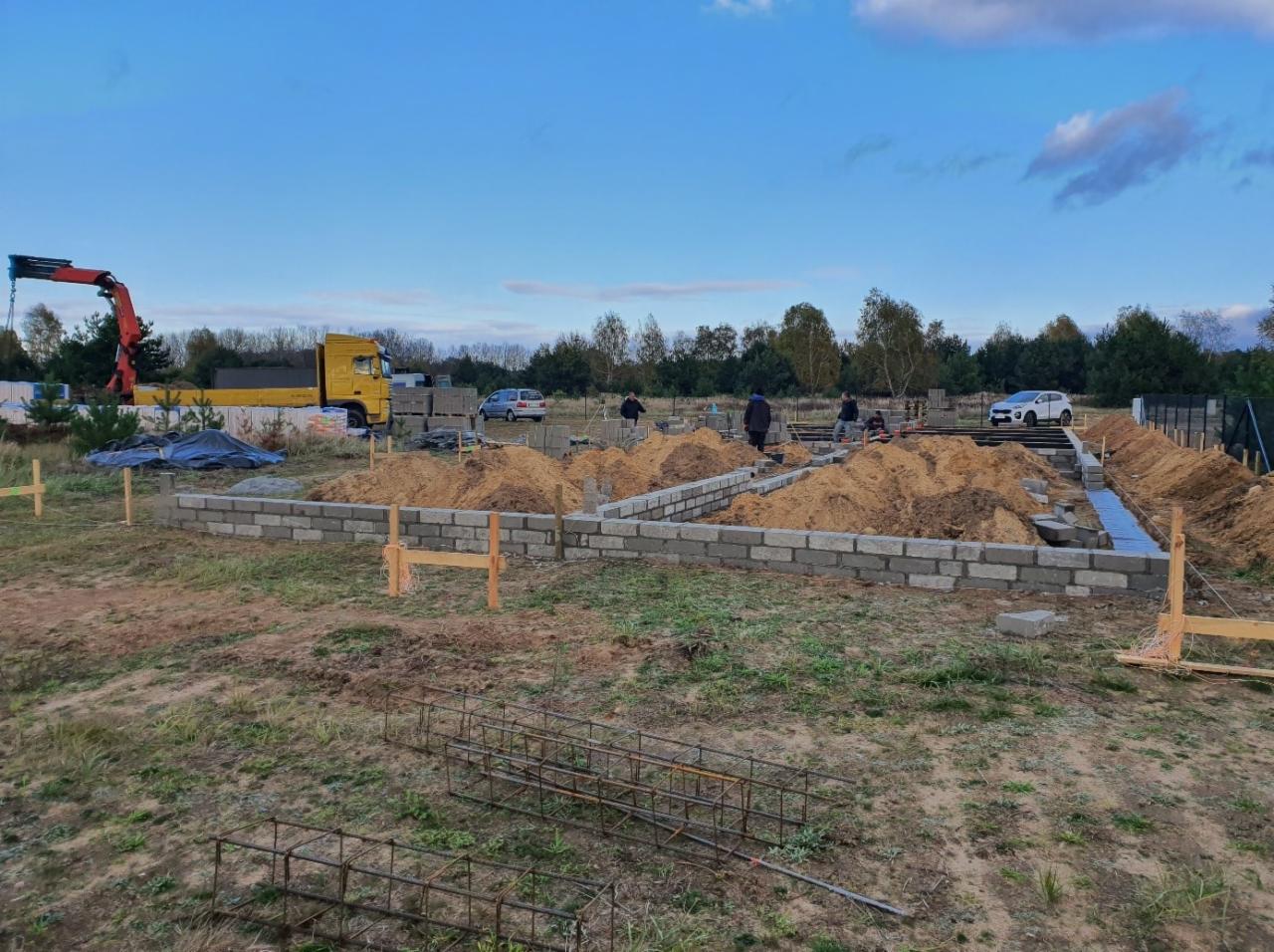 Wznoszenie fundamentów z bloczków betonowych na budowie domu jednorodzinnego, widoczny dźwig samochodowy i zbrojenie w pierwszym planie.