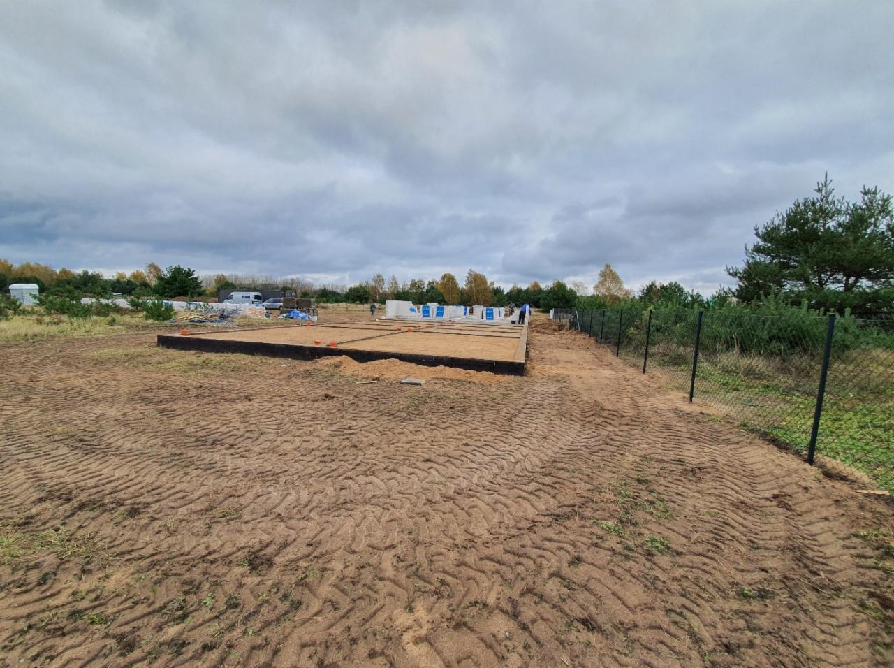 Przygotowany teren pod budowę domu z widocznymi fundamentami w szalunkach, ogrodzenie posesji siatką, w tle drzewa i pochmurne niebo.