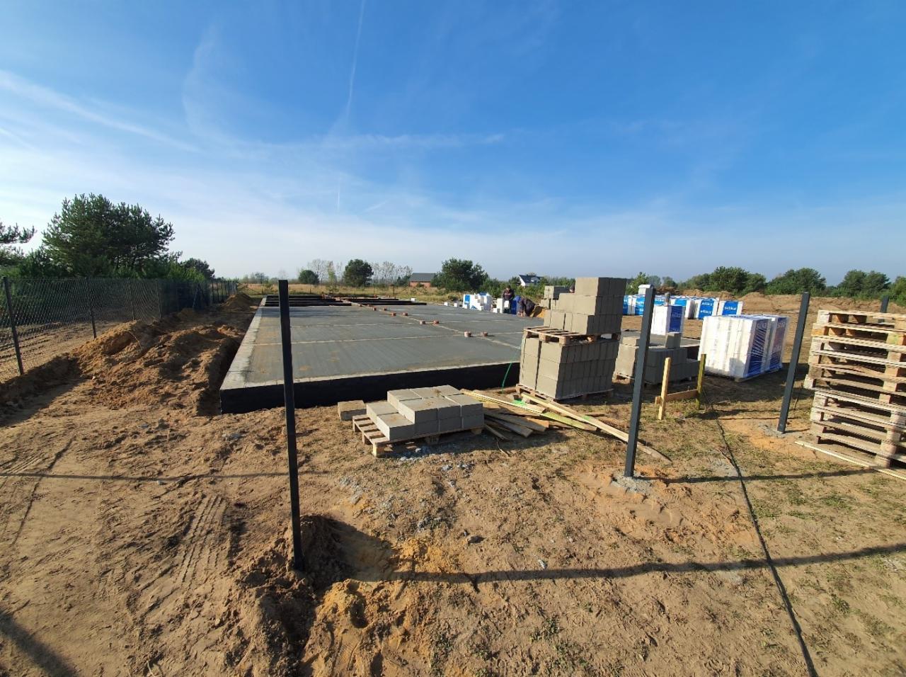 Wylana płyta fundamentowa z izolacją, przygotowana pod budowę domu; na placu budowy palety z bloczkami betonowymi i materiałami izolacyjnymi.