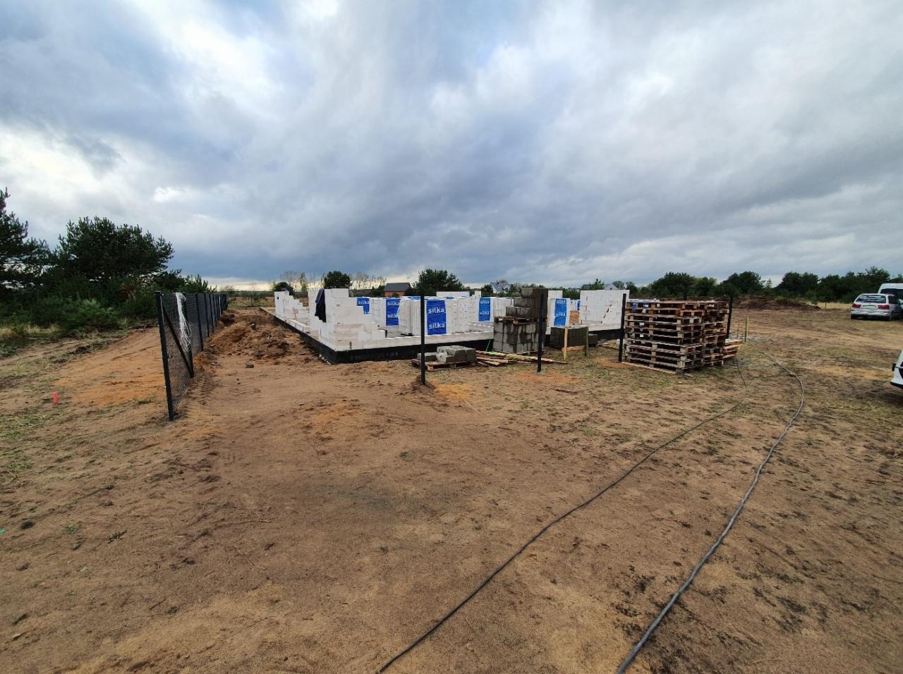 Budowa domu jednorodzinnego w trakcie wznoszenia ścian z bloczków, widoczne fundamenty i palety z materiałami budowlanymi na piaszczystym terenie pod pochmurnym niebem.