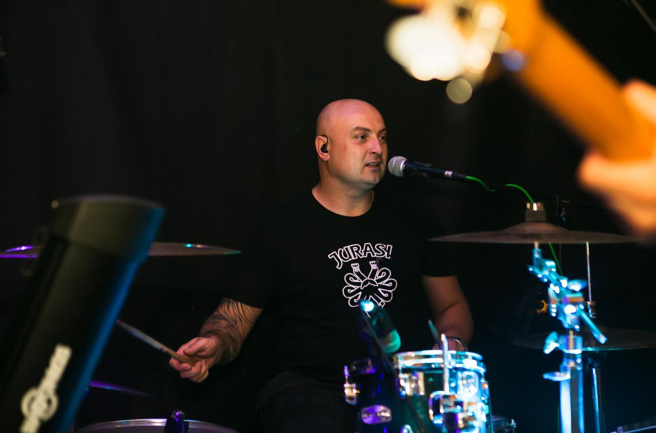 Perkusista z tatuażem na ramieniu gra na perkusji, śpiewając do mikrofonu w trakcie występu na scenie. Na czarnej koszulce widoczny biały nadruk z napisem 'JURASY!'.