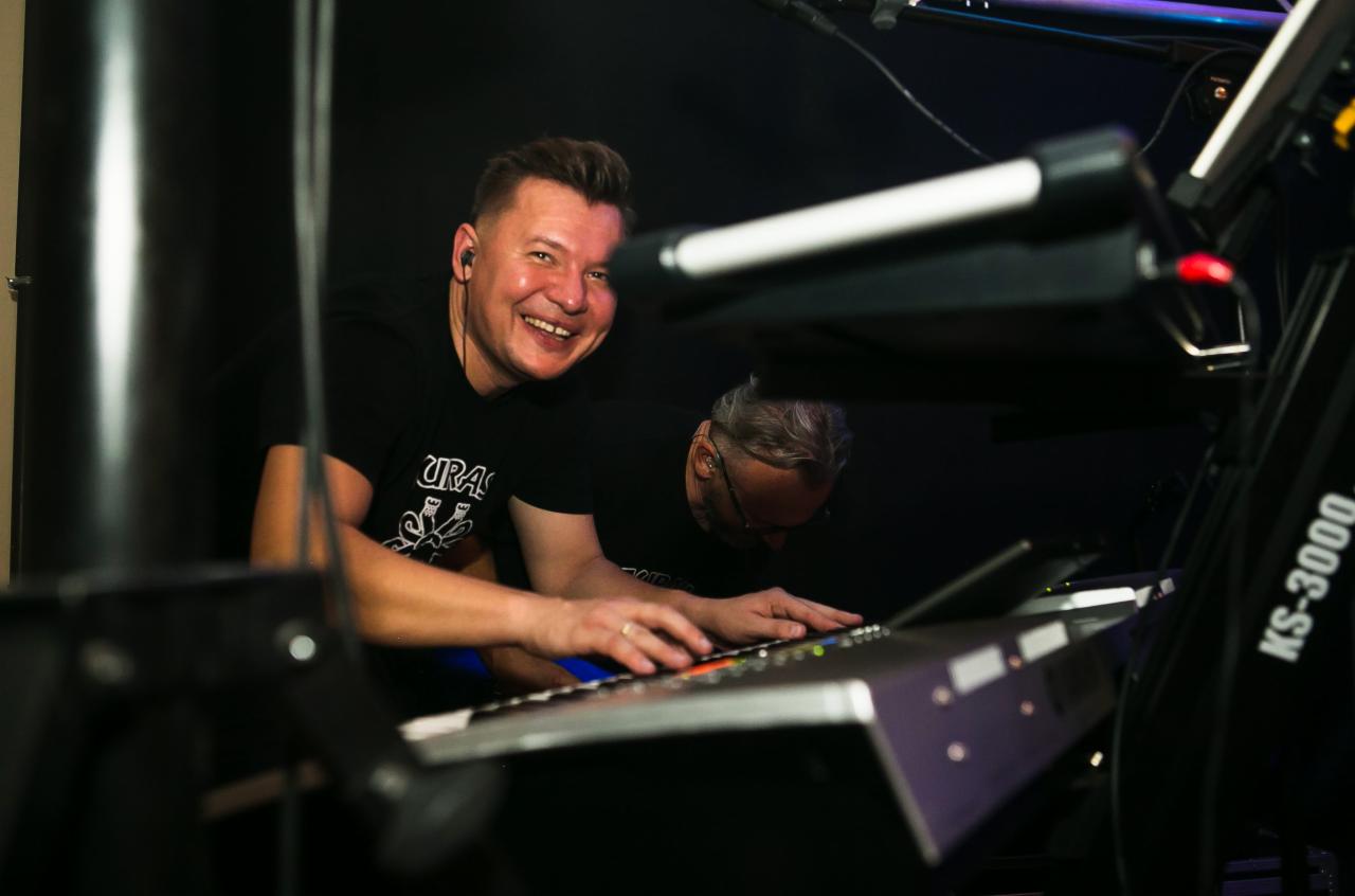 Uśmiechnięty klawiszowiec grający na keyboardzie KS-3000 podczas występu, drugi muzyk w tle skupiony na grze.