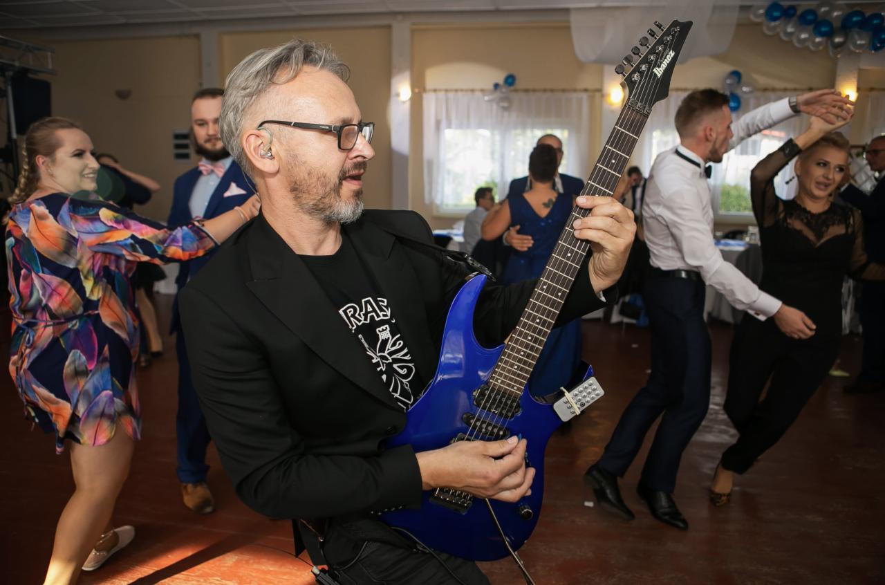Gitarzysta w czarnej marynarce gra na niebieskiej gitarze elektrycznej podczas występu na imprezie, w tle tańczący goście.