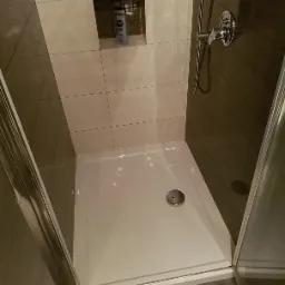 Kabina prysznicowa z brodzikiem, wyłożona beżowymi i brązowymi płytkami, z półką w ścianie, na której stoi butelka kosmetyku. Metalowa armatura i odpływ w brodziku.