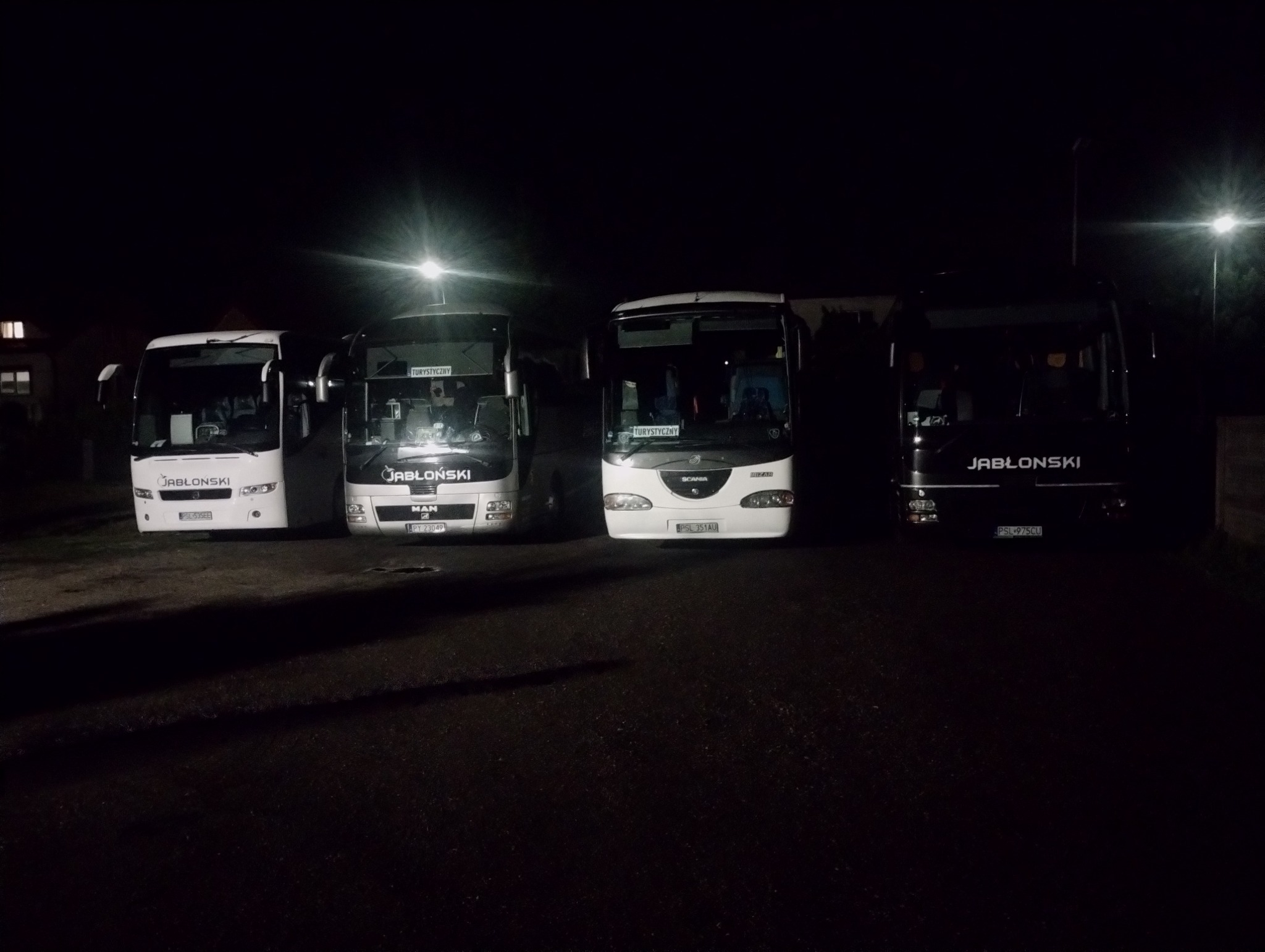 Cztery autokary firmy JABŁONSKI, w tym MAN i SCANIA, zaparkowane nocą pod latarnią. Widoczne tablice 'Turystyczny' i numery rejestracyjne. Ujęcie z dołu.