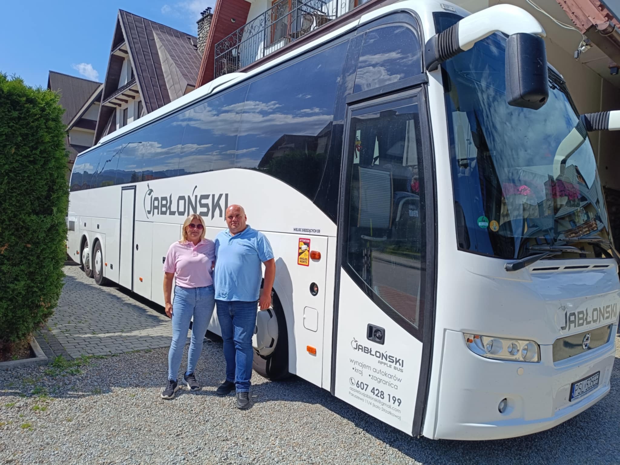 Para stoi przed białym autokarem JABŁOŃSKI Apple Bus z napisem o wynajmie autokarów kraj-zagranica i numerem telefonu, na tle domu z czerwonym dachem.