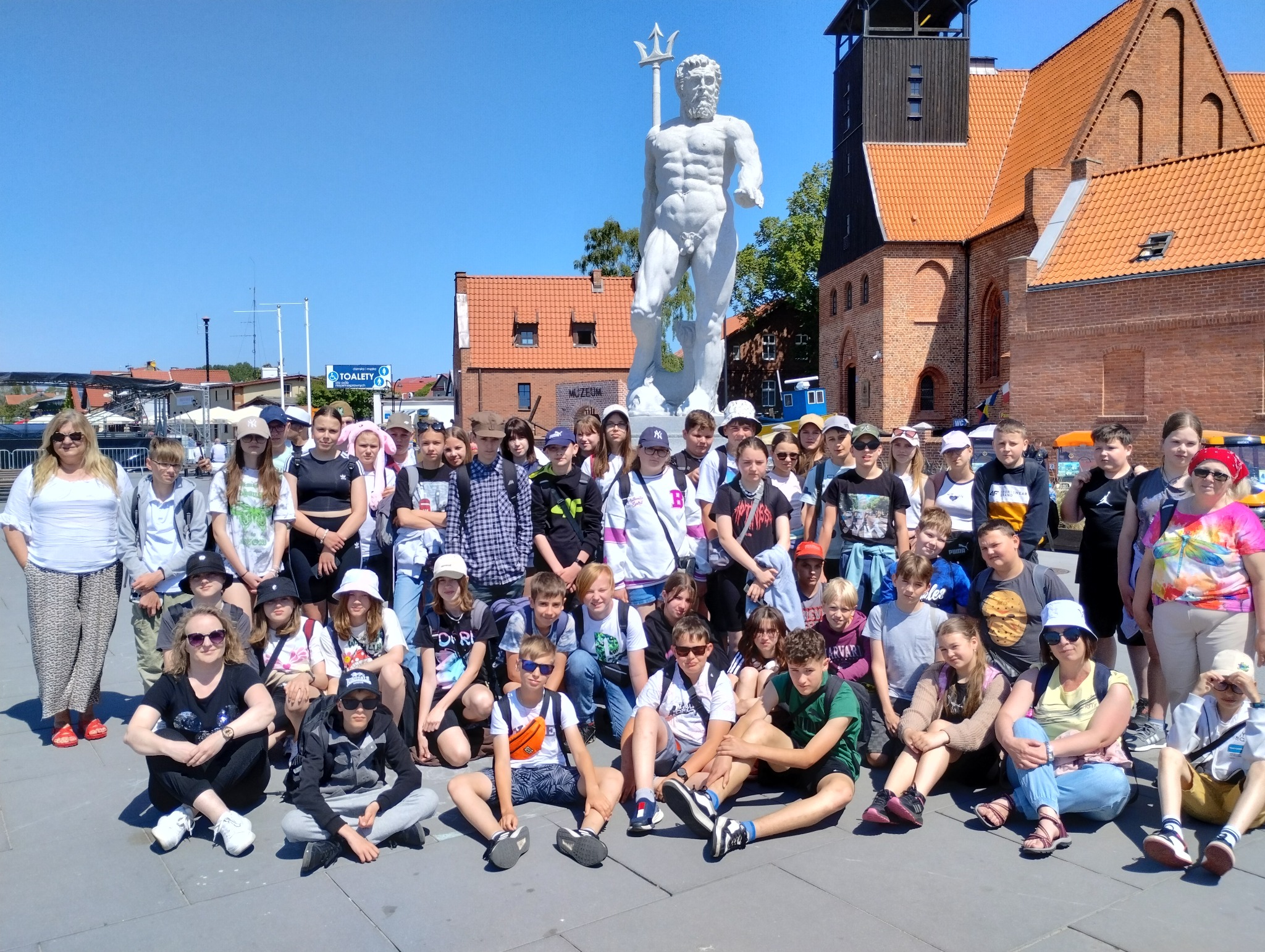 Grupa wycieczkowa pozuje do zdjęcia przed pomnikiem Neptuna w Gdańsku, w tle budynek z czerwonej cegły.