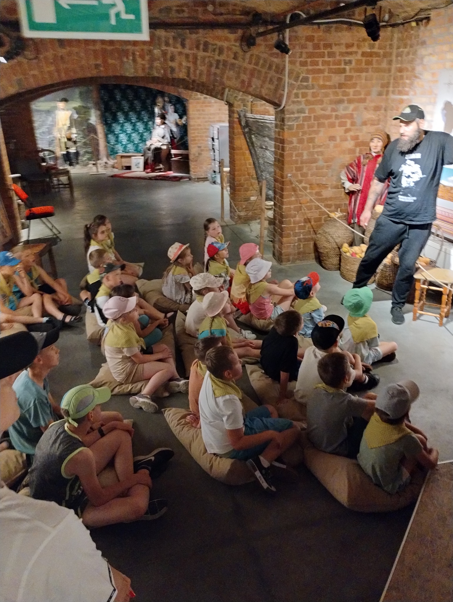 Grupa dzieci siedzi na poduszkach podczas lekcji historii w muzeum, prowadzonej przez przewodnika w czarnej koszulce i czapce, w tle eksponaty i mury z czerwonej cegły.