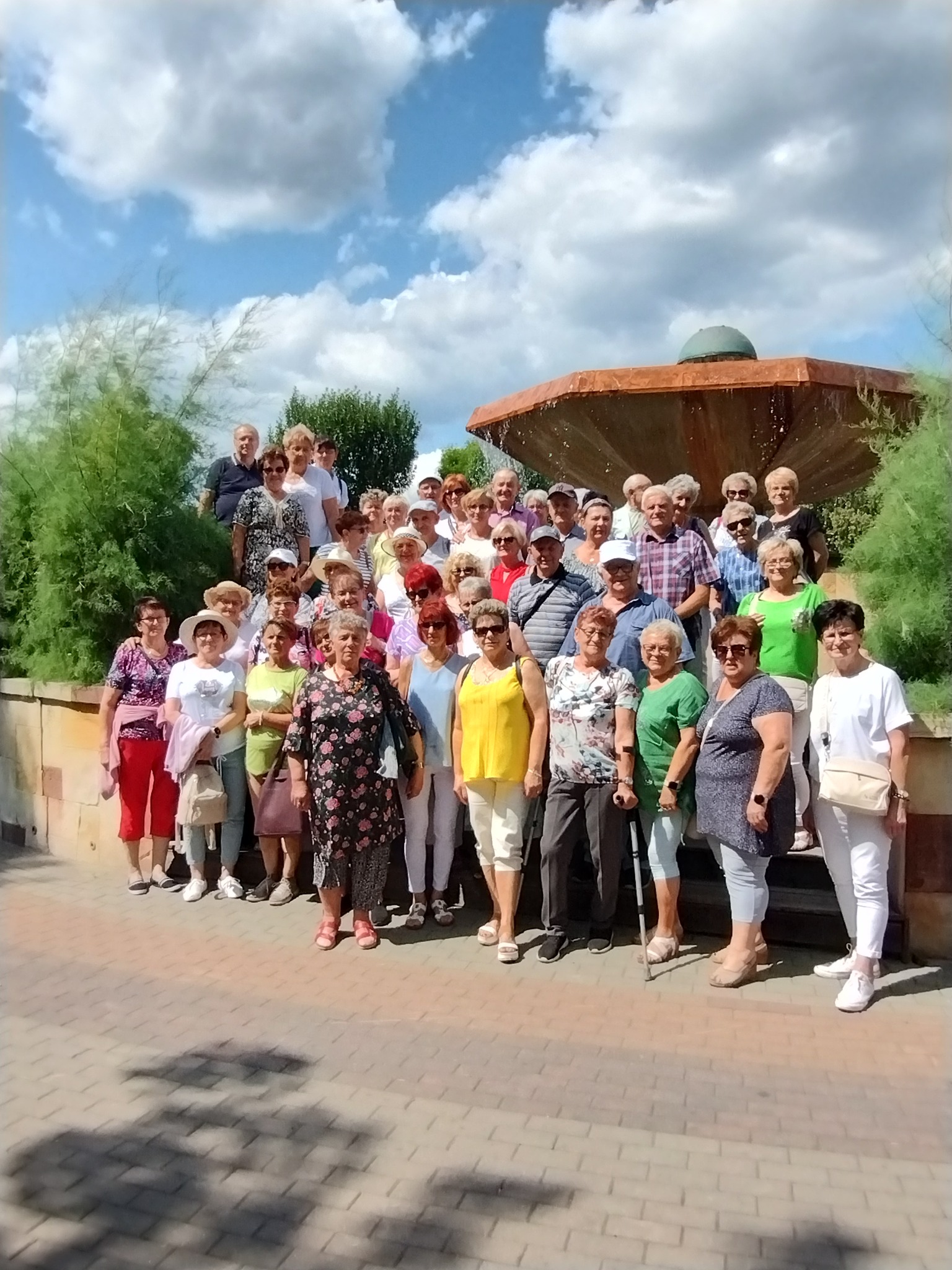 Duża grupa seniorów pozuje do zdjęcia na tle fontanny w słoneczny dzień, część osób w okularach przeciwsłonecznych i kapeluszach.