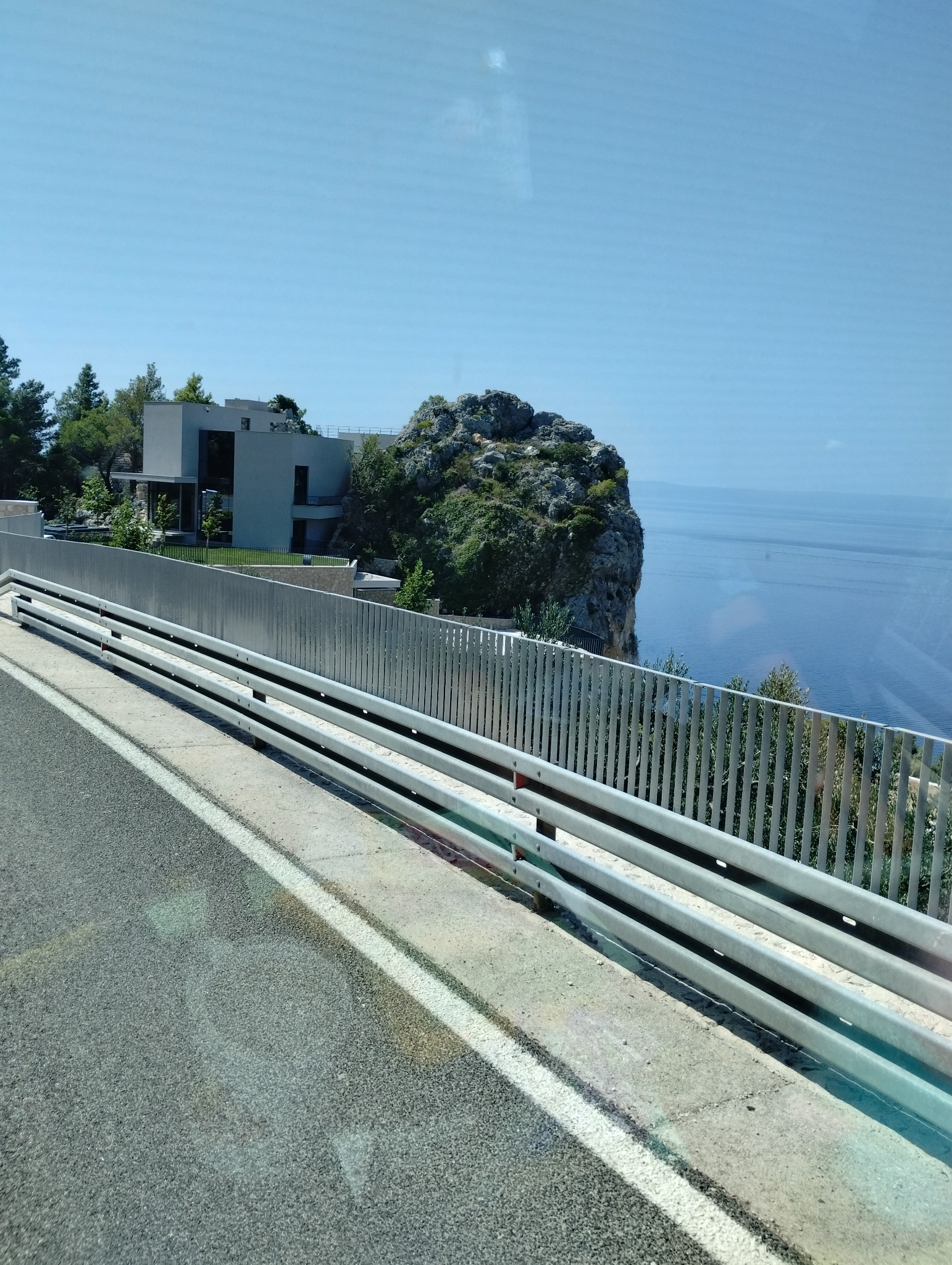 Widok z okna pojazdu na drogę z barierami ochronnymi, nowoczesny dom na skalistym klifie porośniętym zielenią i błękitne morze w tle.