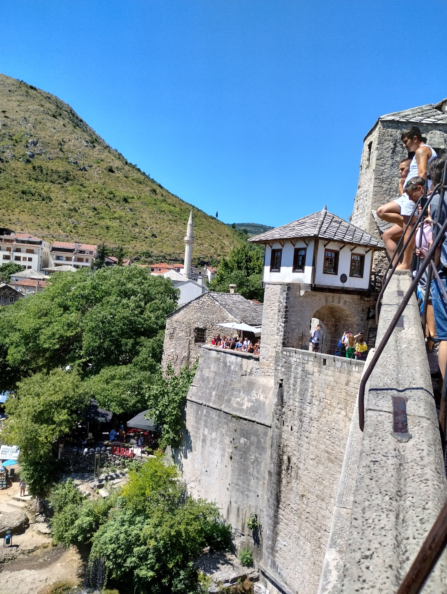 Widok z wysokiego kamiennego mostu na zabytkową architekturę miasta z minaretem i okoliczne wzgórza w słoneczny dzień.