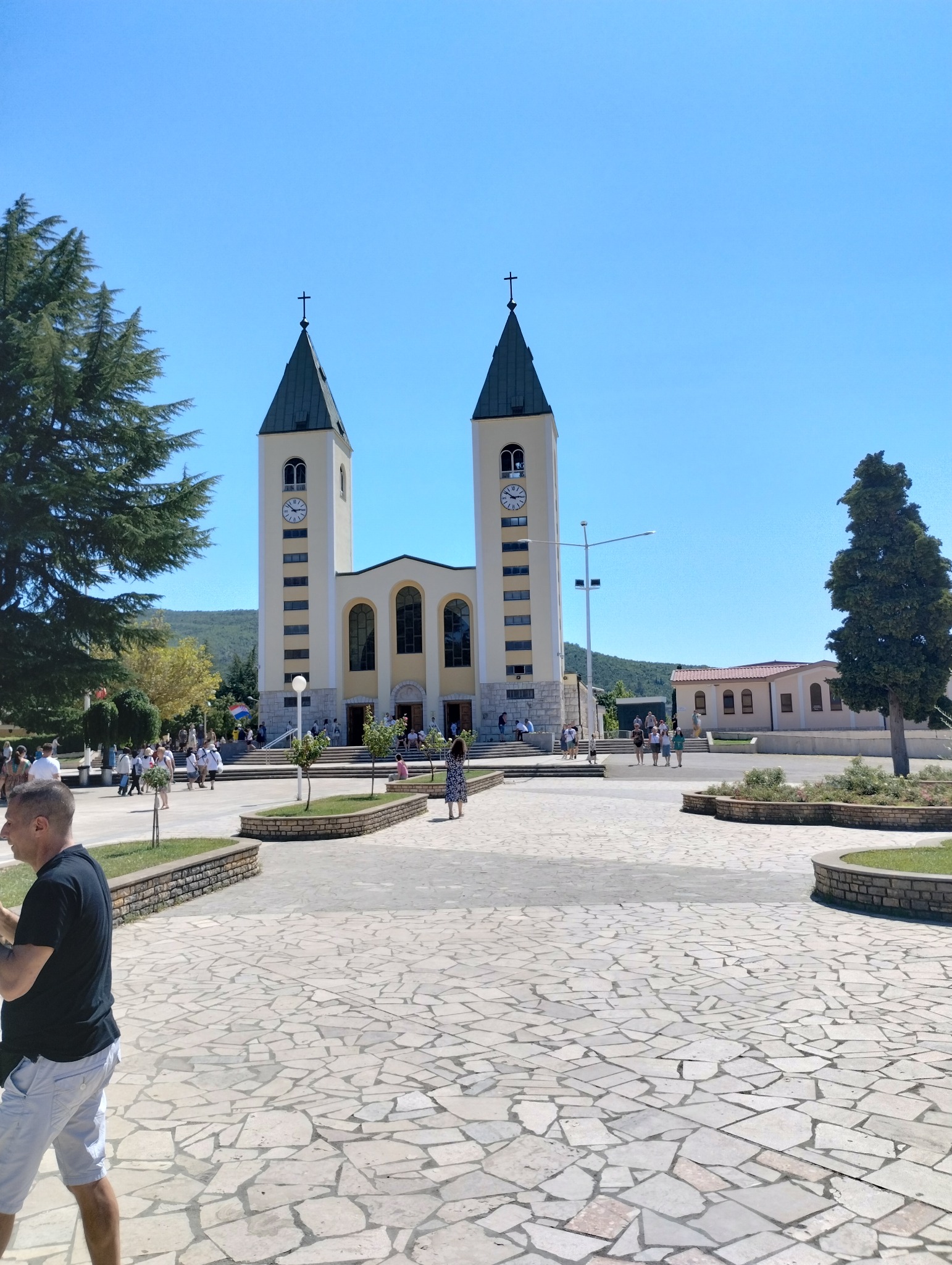 Kościół z dwiema wieżami zegarowymi, plac z kamienną nawierzchnią i spacerujący ludzie w słoneczny dzień.