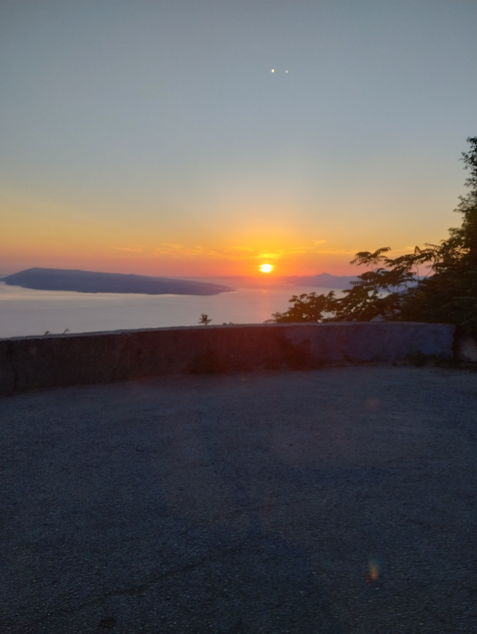 Panoramiczny widok na morze o zachodzie słońca z punktu widokowego, z niskim betonowym murkiem i drzewami po prawej stronie.