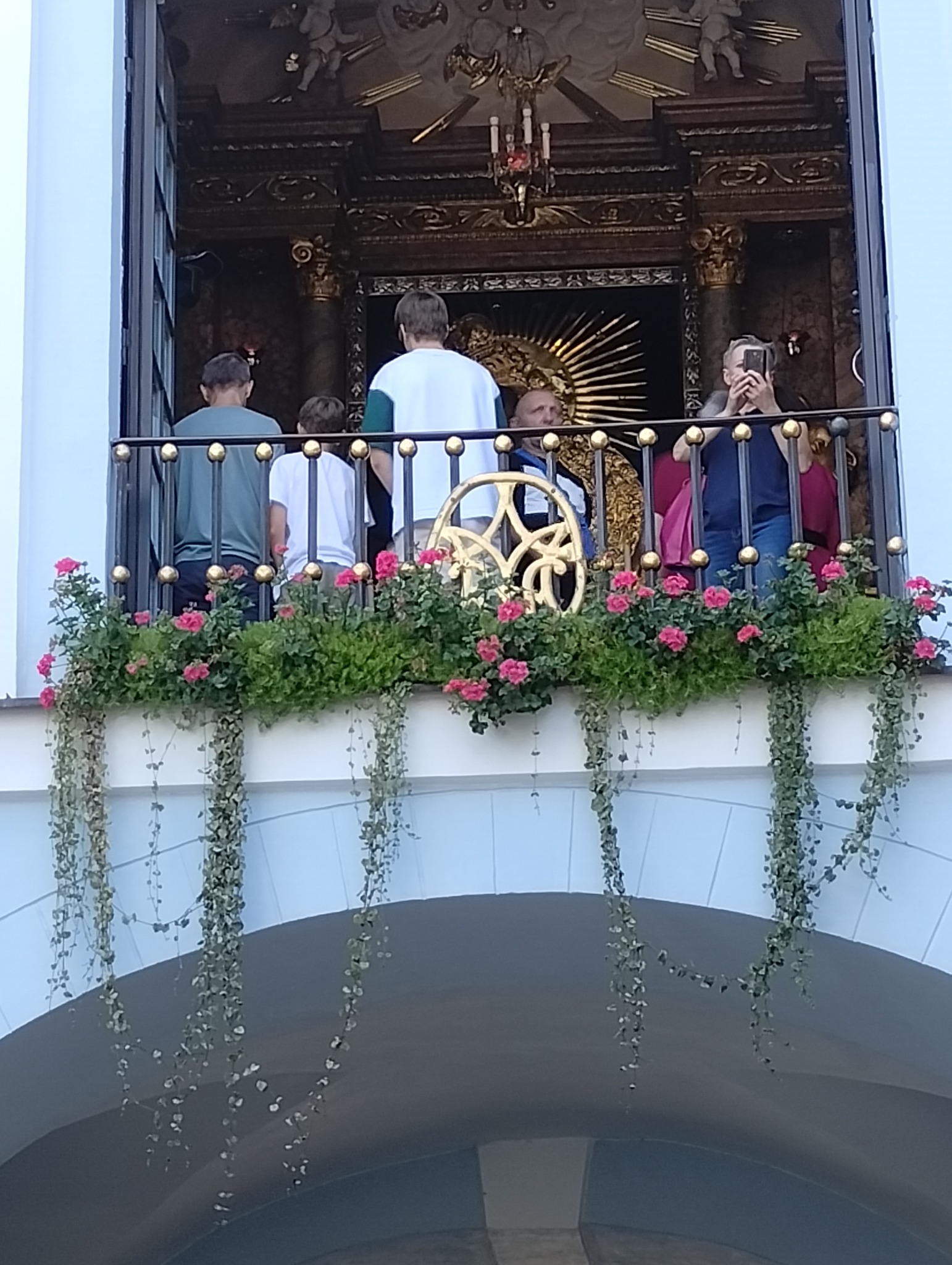 Grupa osób na balkonie z kwiatami, prawdopodobnie podczas zwiedzania zabytkowego obiektu z bogato zdobionym wnętrzem widocznym w tle.