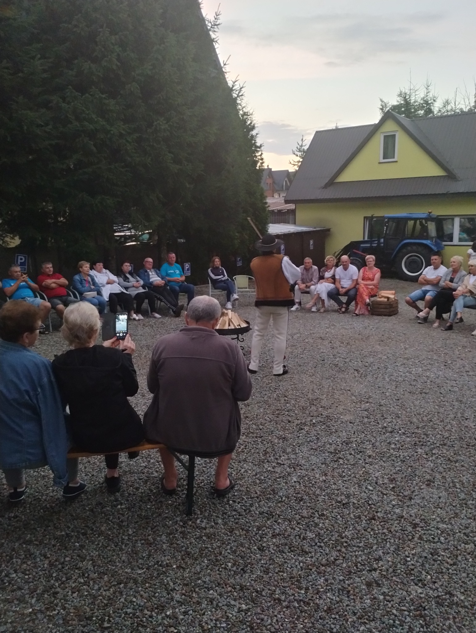 Grupka osób siedzi na zewnątrz, słuchając mężczyzny w stroju regionalnym, który trzyma laskę; w tle budynek i traktor.
