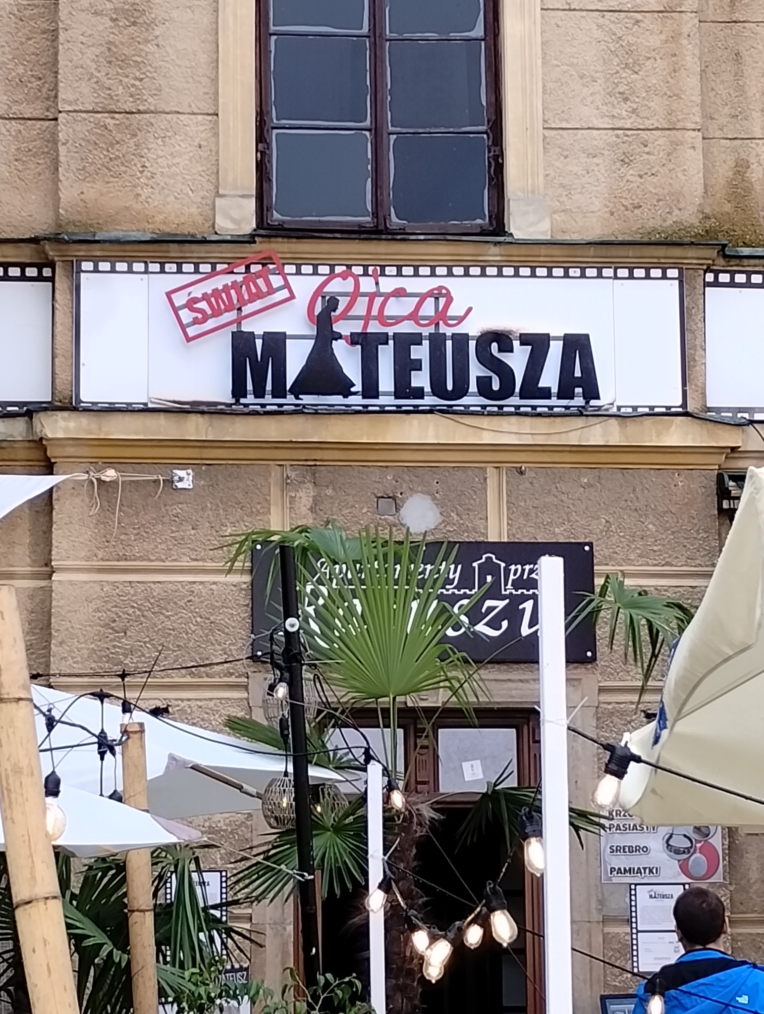 Fasada budynku z szyldem 'Świat Ojca Mateusza', dekoracje z palm i lampek, fragment parasola i sylwetka osoby w niebieskiej kurtce.