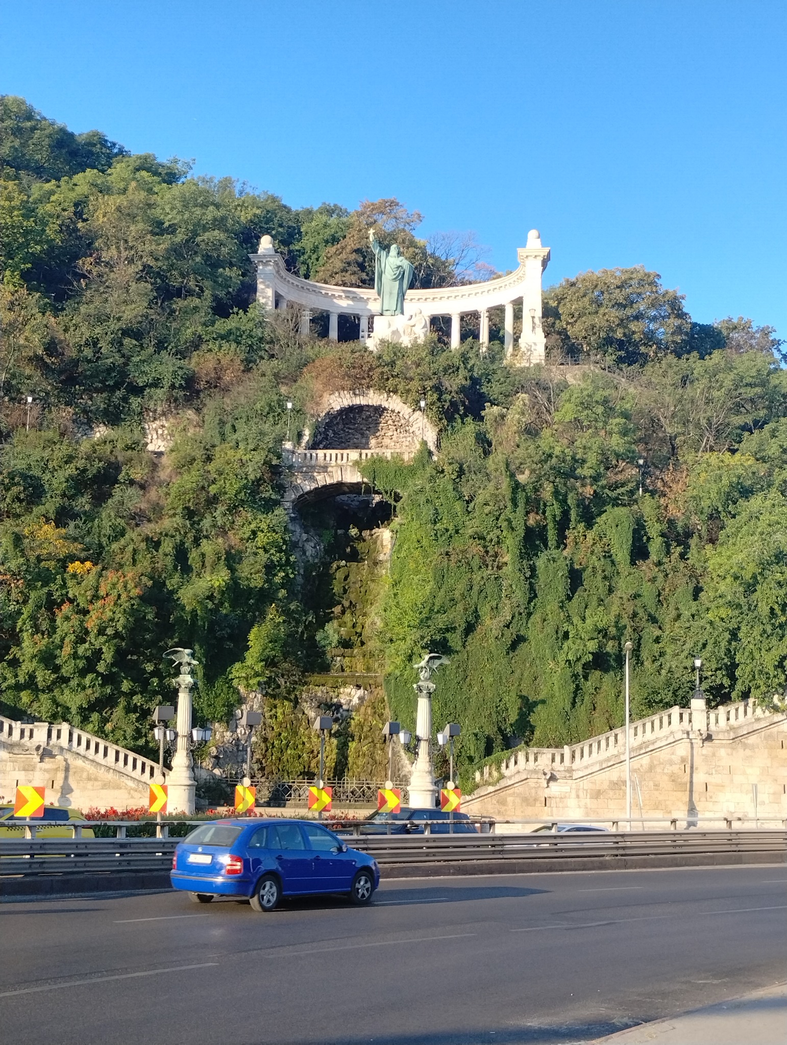 Niebieski samochód osobowy na tle Cytadeli w Budapeszcie, z widoczną statuą Wolności na wzgórzu Gellérta.