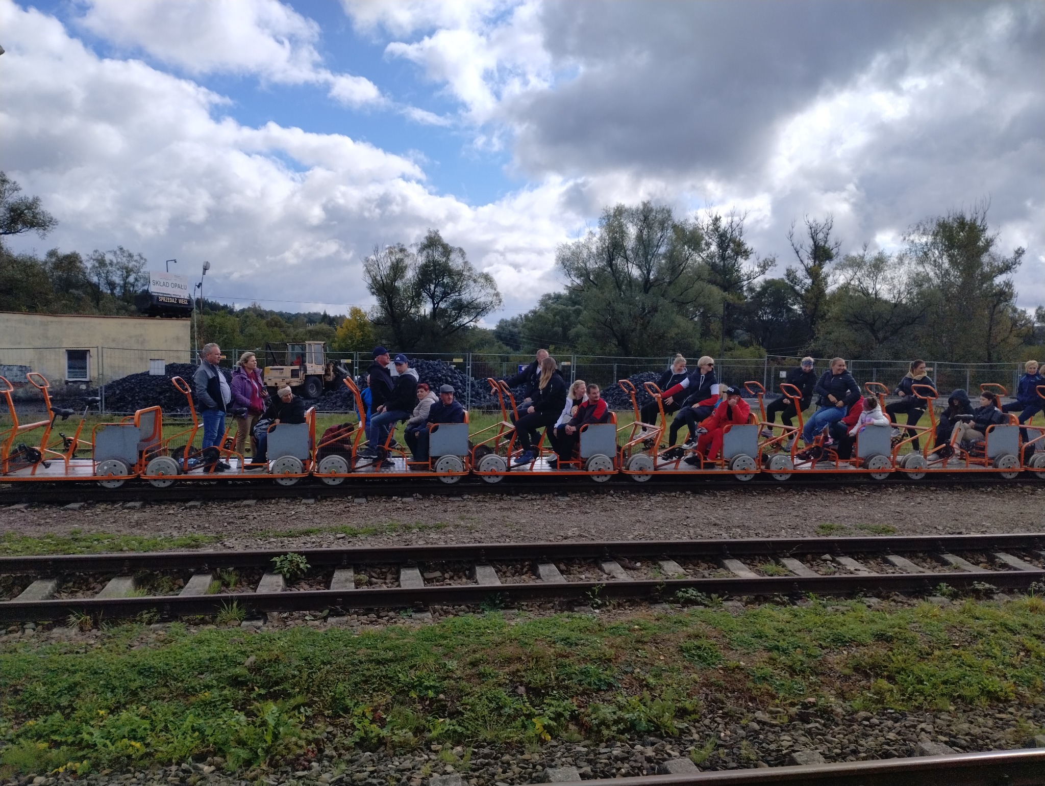 Grupa ludzi na drezynie rowerowej jedzie po torach kolejowych w pochmurny dzień, na drugim planie budynki i hałda węgla.