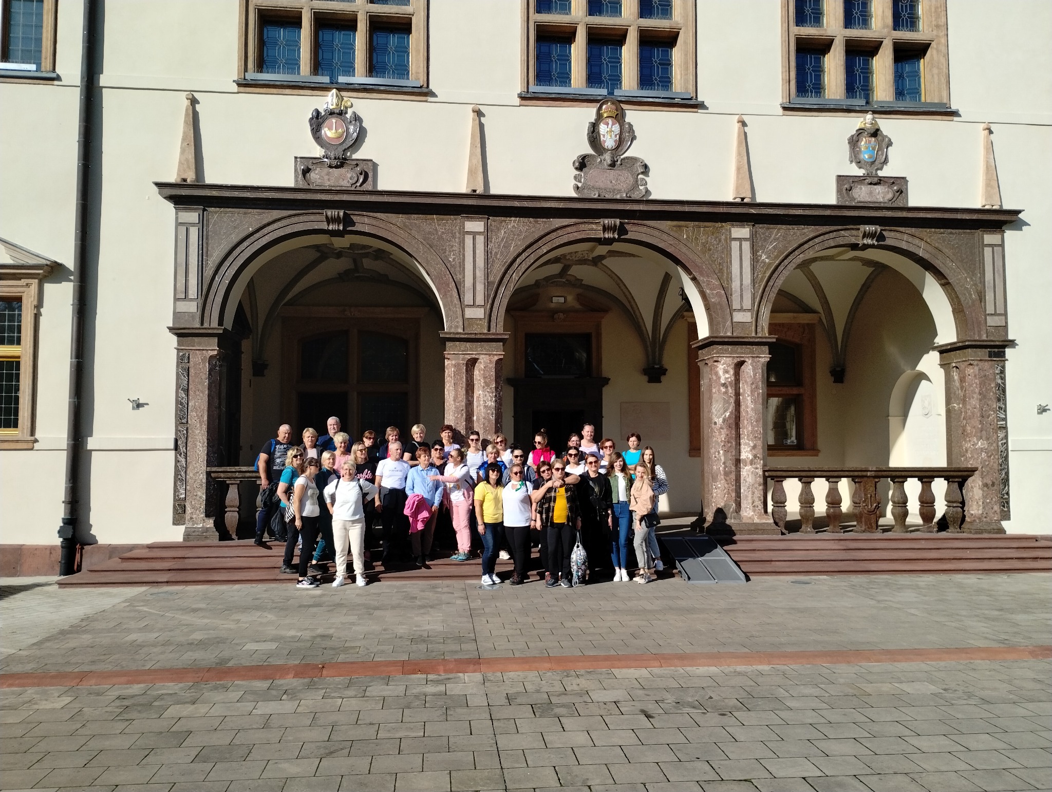 Grupa turystów pozuje na schodach zabytkowego budynku z łukami, widoczny podjazd dla wózków inwalidzkich.