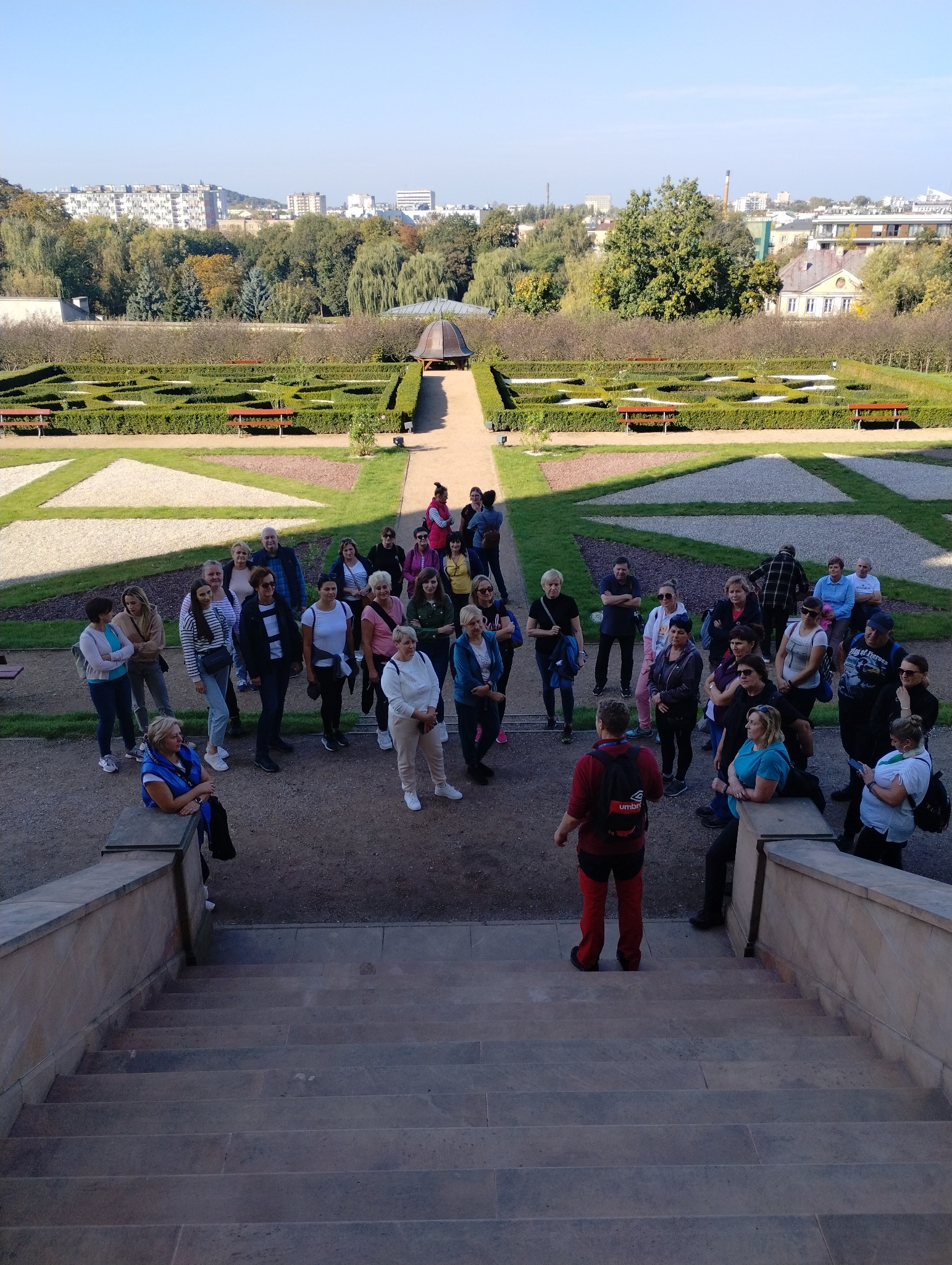 Grupa turystów słucha przewodnika w czerwonym stroju na tle formalnego ogrodu i zabudowy miejskiej.