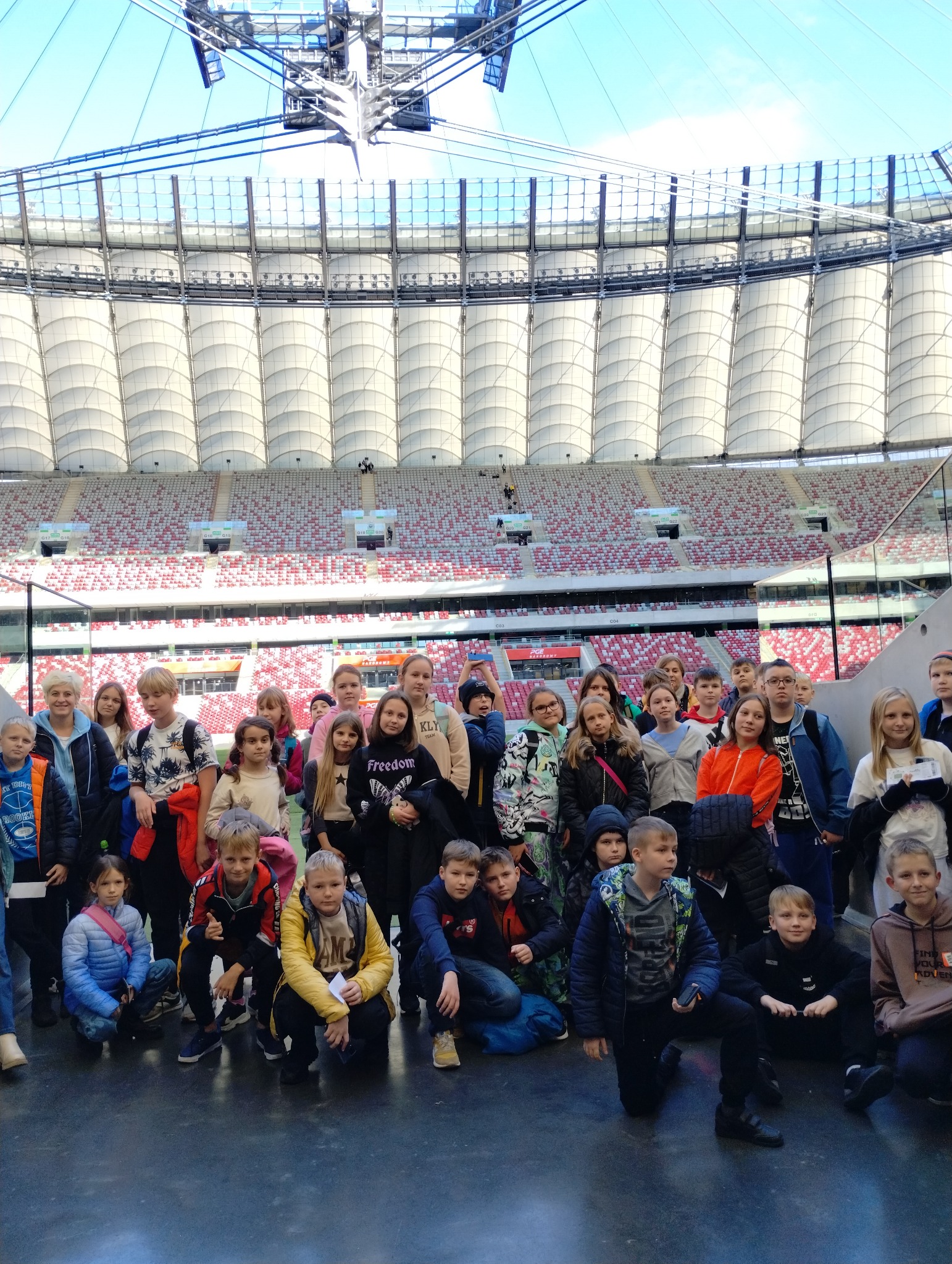 Grupa dzieci i dorosłych pozuje na tle trybun stadionu PGE Narodowy w Warszawie podczas wycieczki.