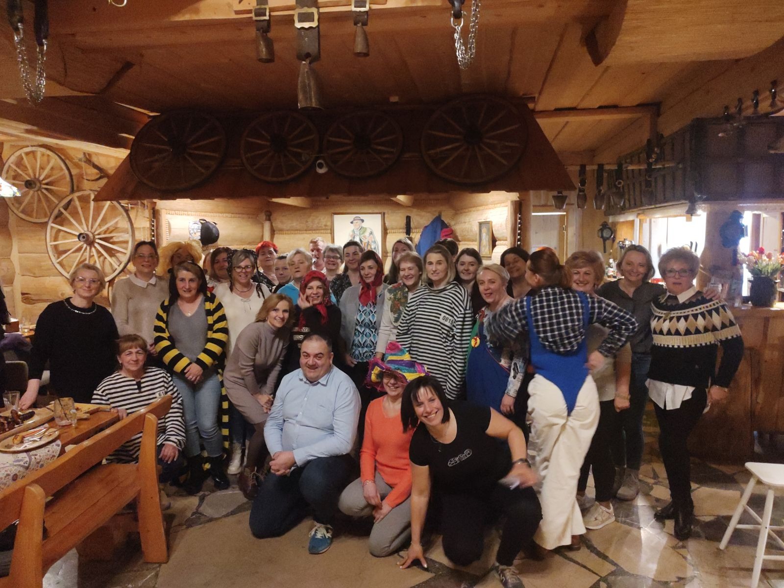 Duża grupa osób pozuje do zdjęcia w drewnianym wnętrzu z kołami wozów na ścianach i dekoracyjnymi dzwonkami pod sufitem.