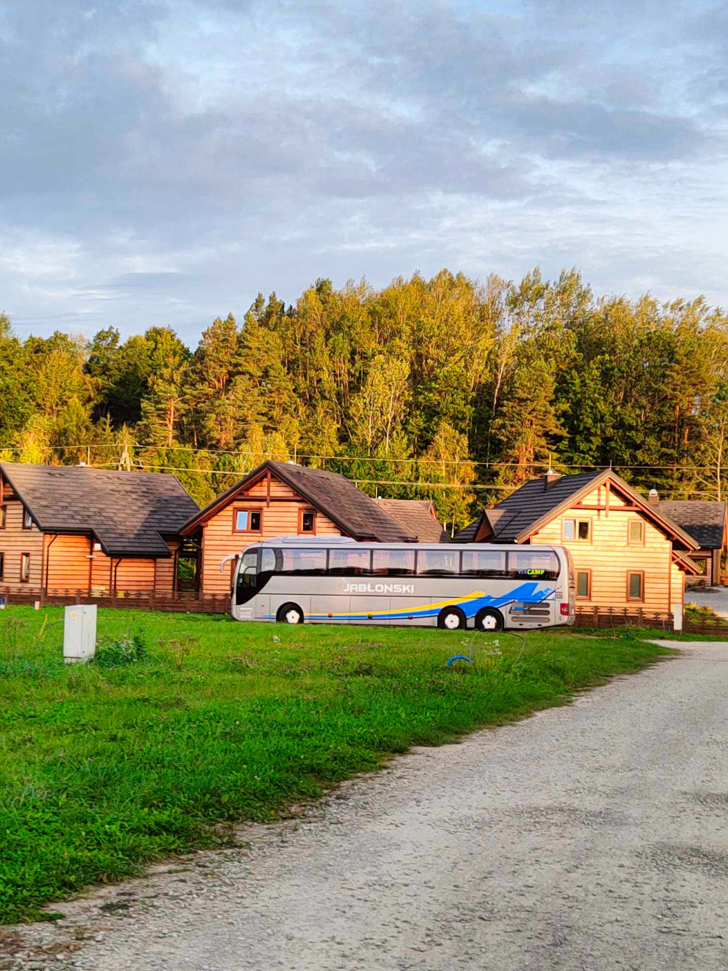 Szary autokar firmy Jabłoński zaparkowany na trawie przed drewnianymi domkami letniskowymi w otoczeniu lasu.