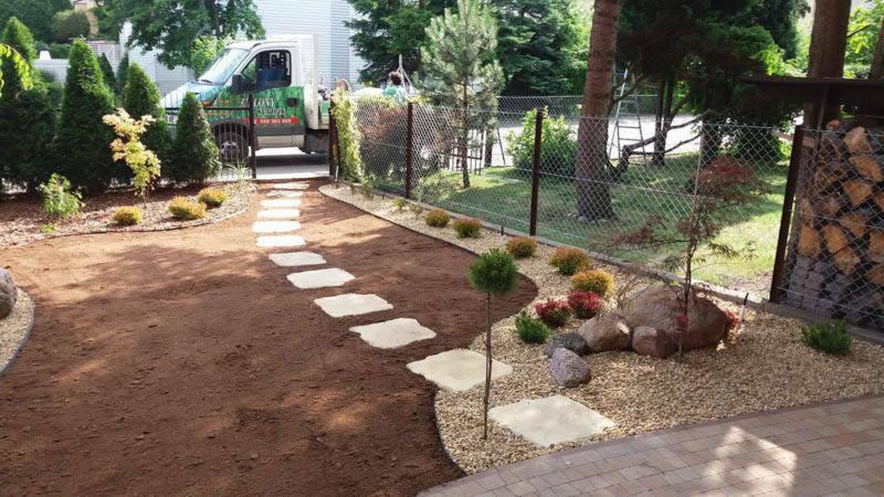 Ścieżka z jasnych płyt prowadząca przez świeżo przygotowany teren pod trawnik, obrzeża obsypane ozdobnym żwirem, w tle widoczny samochód dostawczy z logo firmy ogrodniczej.