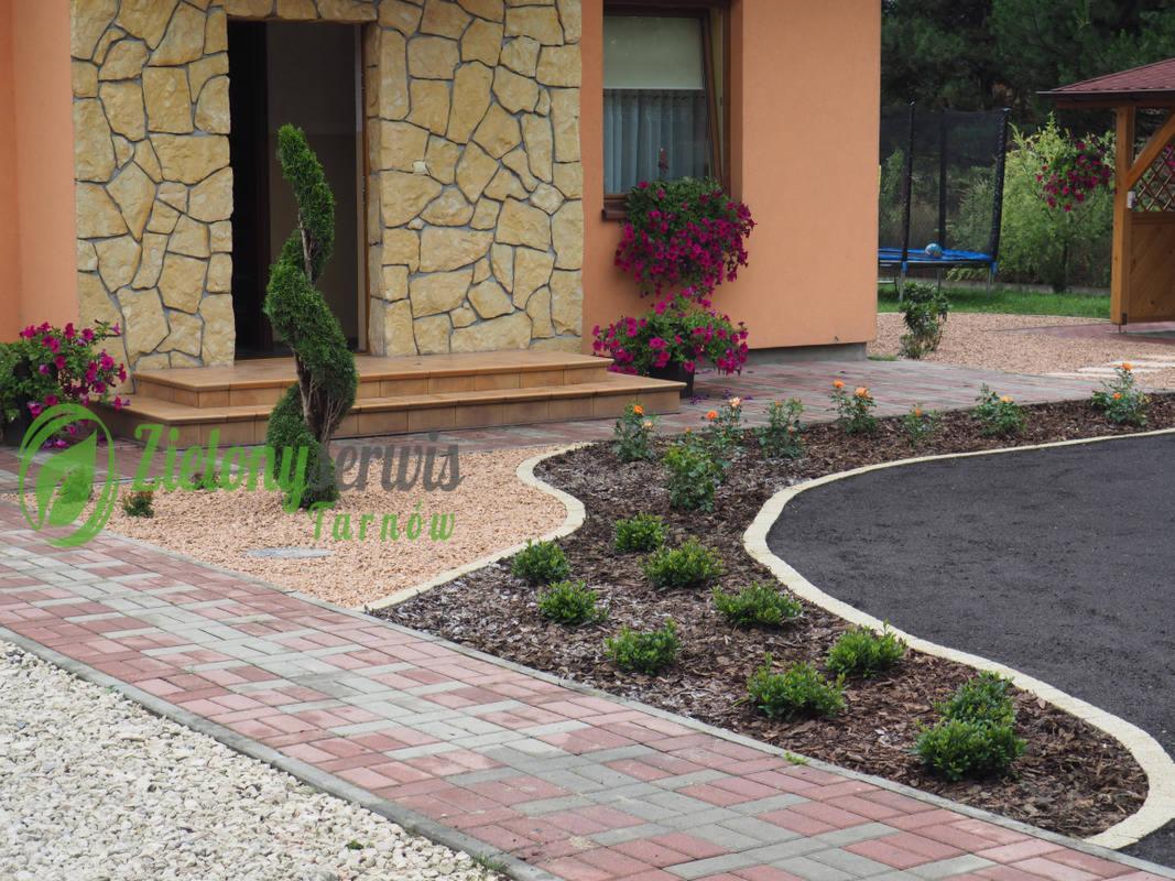 Uporządkowany ogród przydomowy z krętą ścieżką z kostki, rabatami z korą i niskimi krzewami, oraz spiralnie uformowanym drzewkiem iglastym przy wejściu do domu z kamienną elewacją.