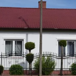 Biały dom z czerwoną dachówką, numerem 30 na elewacji i czarnym, ozdobnym ogrodzeniem z topiarami przed oknami.