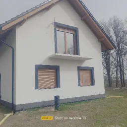 Dom jednorodzinny z nową, jasną elewacją, szarą stolarką okienną i drewnianym wykończeniem dachu, widok z boku na tle drzew bez liści.