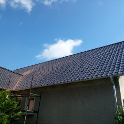 Jar-Bud - Ciemna dachówka ceramiczna na skośnym dachu domu jednorodzinnego, widok z dołu, fragment rusztowania, błękitne niebo z chmurami w tle.