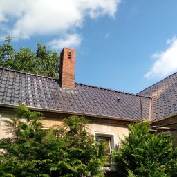 Jar-Bud - Błyszczący, ciemny dach z dachówki ceramicznej na tle błękitnego nieba z chmurami. Widoczny komin z cegły i fragment elewacji budynku z zielenią.