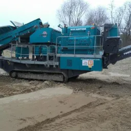 Niebieska przesiewarka mobilna 1065 MAXTRAK na gąsienicach, stojąca na piaszczystym terenie z widocznymi śladami opon, w tle drzewa bez liści pod pochmurnym niebem.