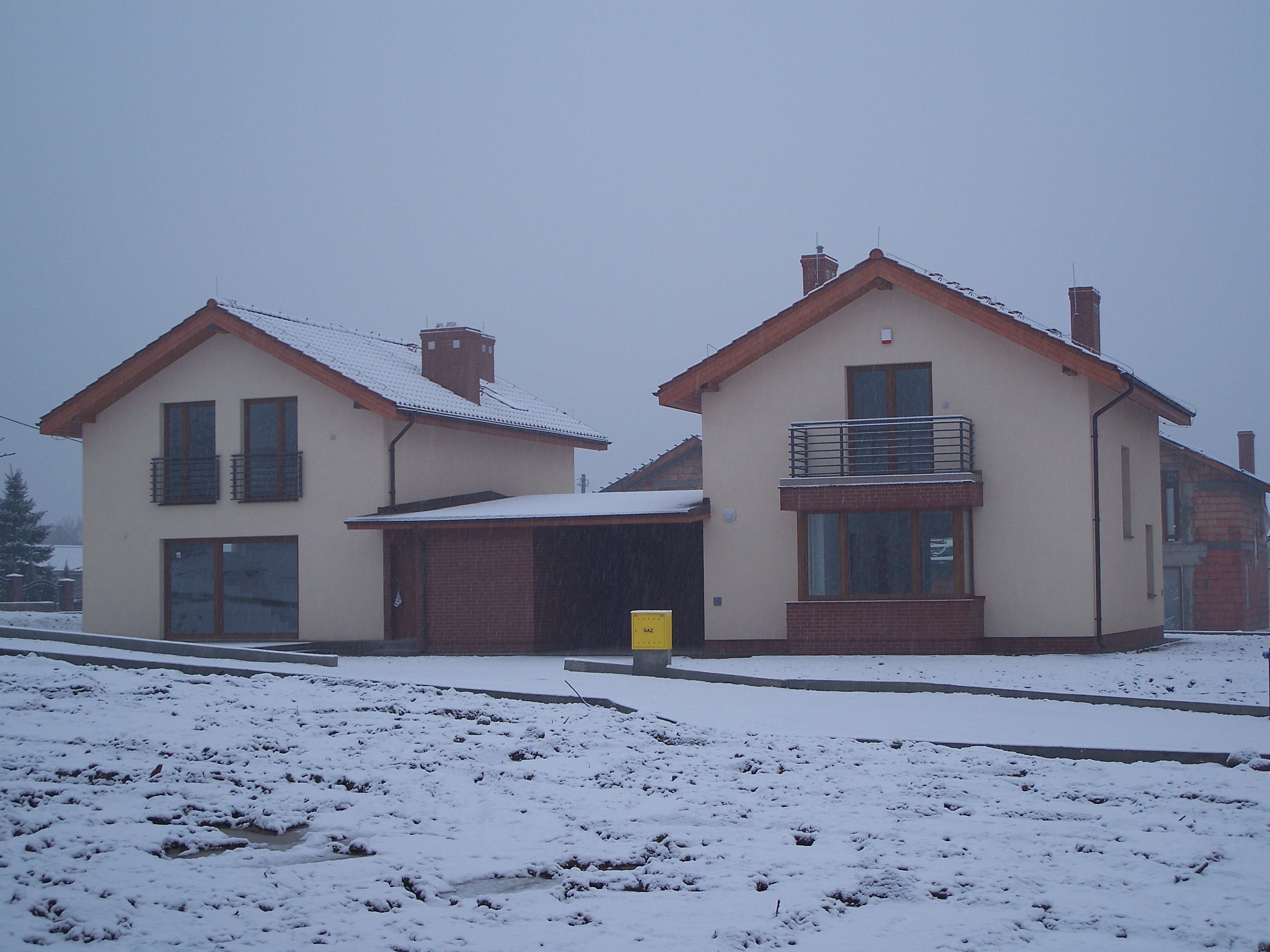 Dwa nowe domy jednorodzinne w trakcie budowy, z widocznymi niezakończonymi elewacjami i zasypanym śniegiem terenem wokół, w pochmurny zimowy dzień.