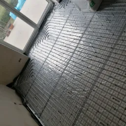 Układanie rur grzewczych na macie izolacyjnej z widocznymi napisami SCHILTE, przygotowanie do wylewki pod ogrzewanie podłogowe w nowym budynku, widoczne okno i materiały izolacyjne XPS.