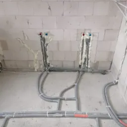 Surowe wnętrze w trakcie prac hydraulicznych: szare rury instalacji wodnej wystające ze ściany z betonu komórkowego, zabezpieczone pianką montażową, widoczne łączenia rur.