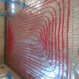 Czerwone rury ogrzewania podłogowego przymocowane do siatki na ścianie, widoczne niebieskie rury instalacji wodnej w górnym rogu.