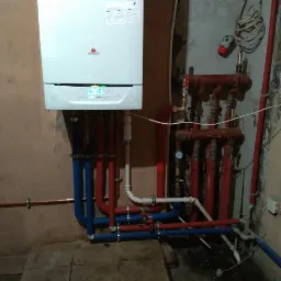 Biały piec gazowy zamontowany na ścianie z podłączonymi rurami w kolorach niebieskim i czerwonym. Obok widoczny rozdzielacz z licznymi zaworami i rurami. Tło stanowi surowa ściana z widocznymi...