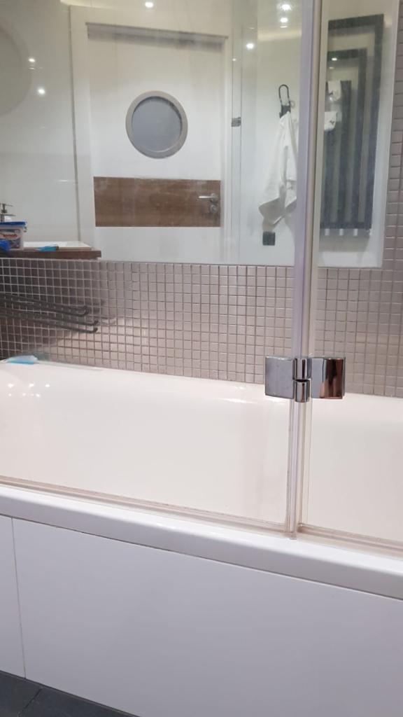 Szklana kabina prysznicowa zamontowana na wannie, widoczna mozaika na ścianie i okrągłe okno w drzwiach łazienki w tle.