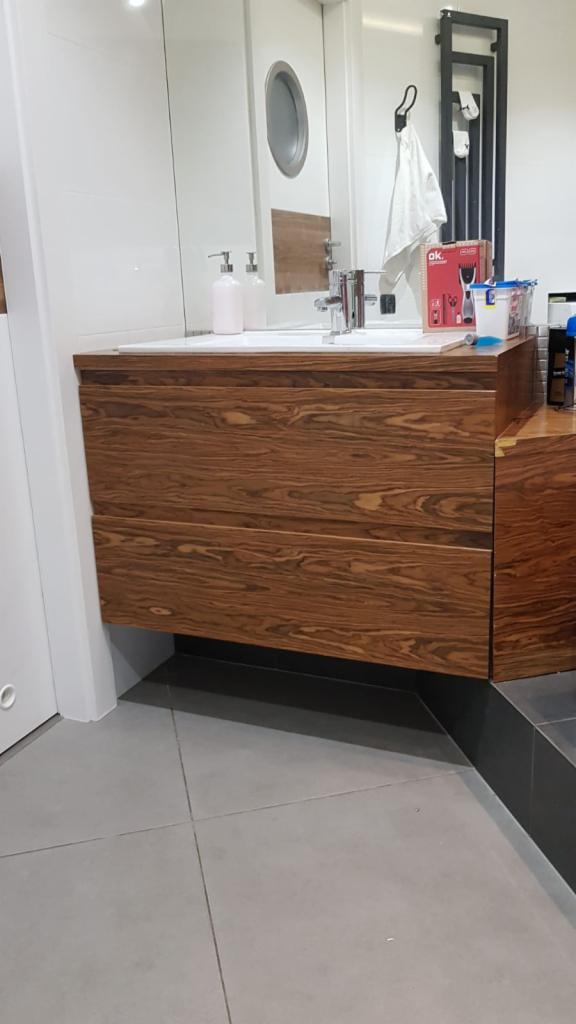 Nowoczesna szafka pod umywalkę z forniru o wyraźnym usłojeniu w łazience z szarą podłogą i białymi płytkami, widoczne akcesoria łazienkowe na blacie.