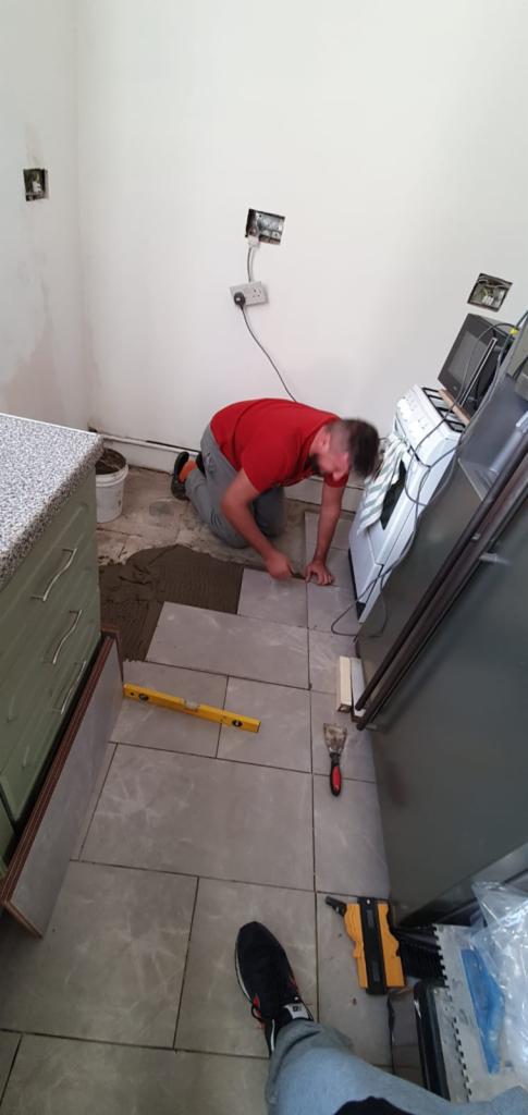 Mężczyzna w czerwonej koszulce docina płytki podłogowe w kuchni, widoczny poziomica, szpachelka i klej w wiadrze. Częściowo ułożona podłoga z szarych płytek.